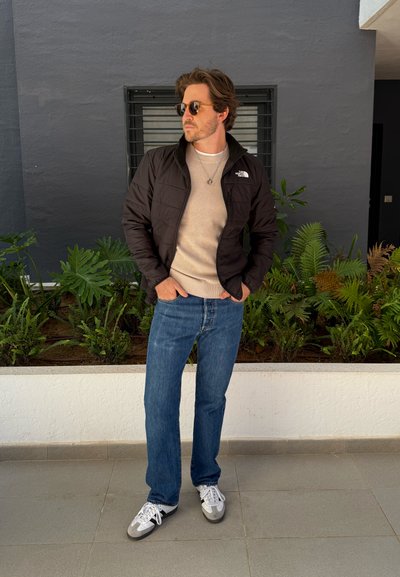 Chaqueta negra aislante, suéter beige acanalado, jeans azules y zapatillas grises. De pie entre vegetación con una pared gris de fondo.