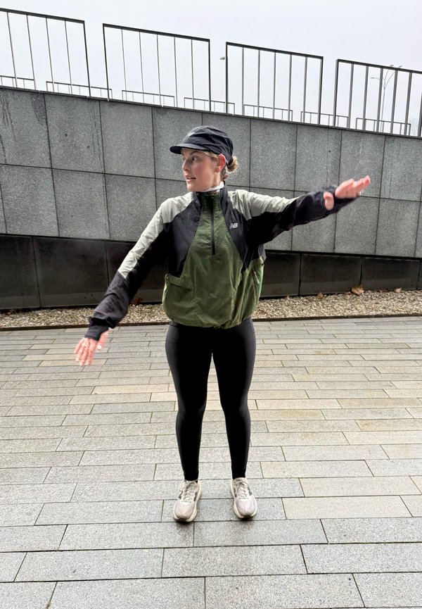 Green and black windcheater with a zip-up front, long sleeves, and an elasticated hem, paired with black leggings and grey trainers on a paved surface.