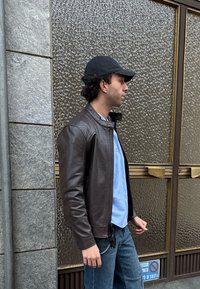 Άνδρας με μαύρο καπέλο, καφέ δερμάτινο μπουφάν, μπλε πουκάμισο και τζιν περπατάει μπροστά από υαλοπίνακες με υφή, ενώ υπάρχει σήμα μη στάθμευσης στο πεζοδρόμιο.