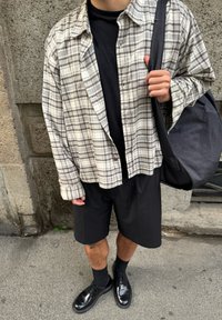 Personne portant un t-shirt noir, un short noir, des chaussures noires avec des chaussettes, et une chemise à carreaux beige, tenant un sac à bandoulière noir contre un mur de pierre.