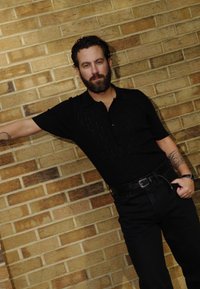 Μαύρο πόλο πουκάμισο κοντό μανίκι με κατακόρυφο υφασμάτινο σχέδιο, συνδυασμένο με ψηλόμεσα μαύρα τζιν. Στηρίζεται σε έναν τοίχο από τούβλα.