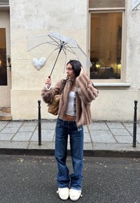 Jeune femme en veste en fourrure tenant un parapluie transparent dans une rue pluvieuse, avec un bâtiment beige et une décoration en forme de cœur en arrière-plan.