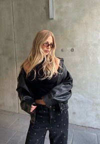Chaqueta de cuero negra de gran tamaño llevada sobre una camiseta negra ajustada, combinada con jeans negros con detalles de ojales de metal. Las gafas son rectangulares y tintadas.