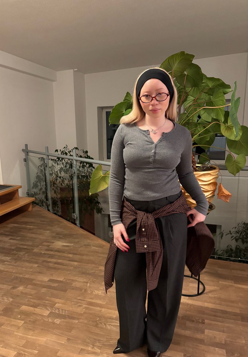 Gray long-sleeve shirt with button details, black wide-legged pants, and a brown plaid shirt tied at the waist. Black headband and glasses.