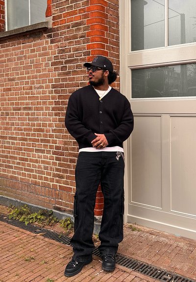 Cardigan negro, camiseta blanca, pantalones cargo negros con bolsillos laterales y deportivas oscuras. Gafas de sol cuadradas y una gorra negra completan el look.