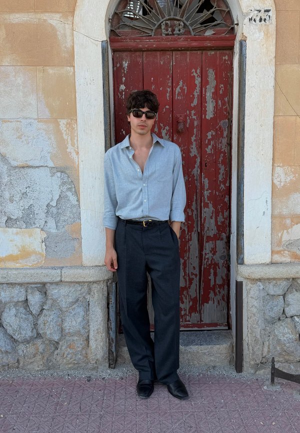 Chemise en lin bleu clair, pantalon noir et chaussures noires. Debout contre une porte en bois rouge vieilli avec de la peinture écaillée et un mur en pierre.