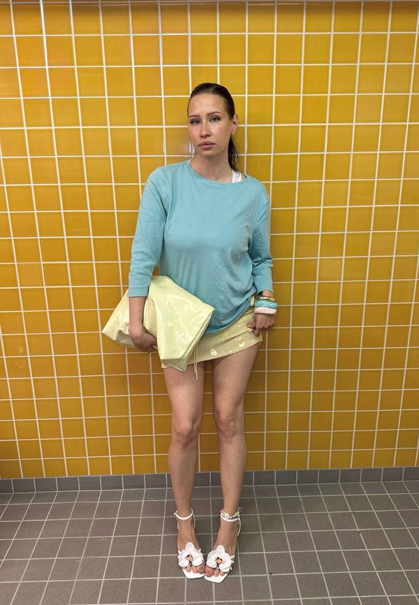 Vrouw met een blauw hemd met lange mouwen, gouden minirok, witte sandalen met bloemen, die een grote gele tas vasthoudt tegen een gele betegelde muur.