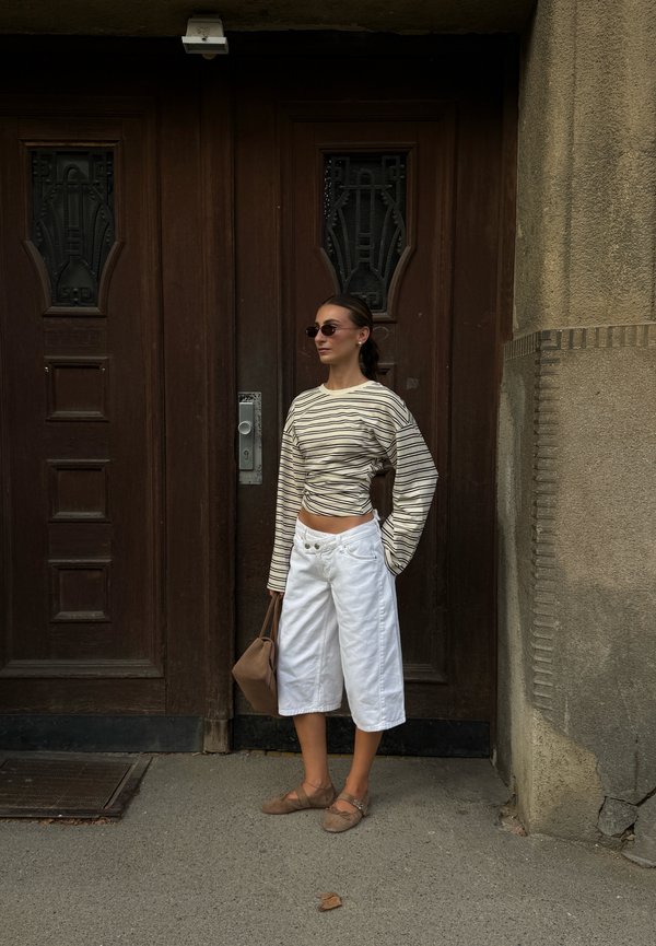 Femme portant des lunettes de soleil, un t-shirt rayé à manches longues, un short blanc large et des ballerines marron, se tient devant une porte en bois en tenant un sac marron.