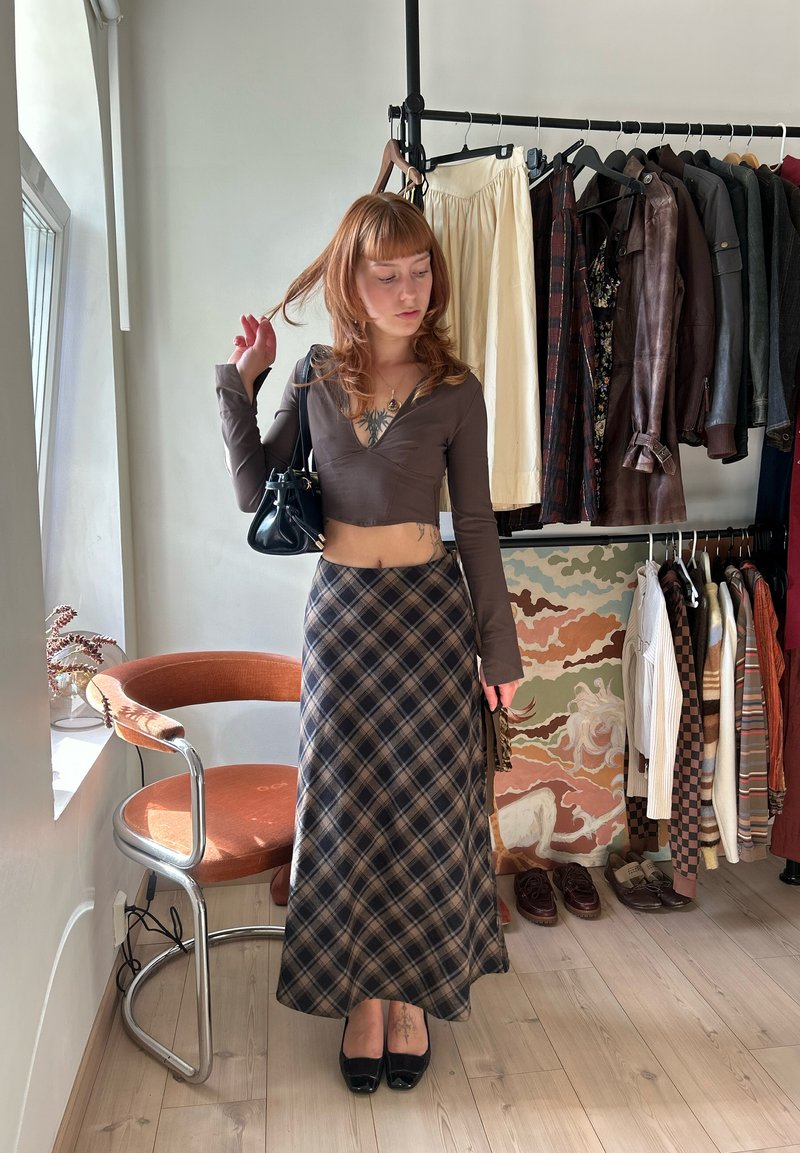 Jeune femme aux cheveux roux portant un crop top marron et une longue jupe à carreaux, debout dans une pièce avec des vêtements suspendus sur des portants derrière elle.