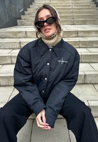 Fekete párnázott dzseki nagy gallérral, ezüst gombokkal és "Calvin Klein Jeans" logóval; világos színű kötött sállal és fekete nadrággal párosítva.