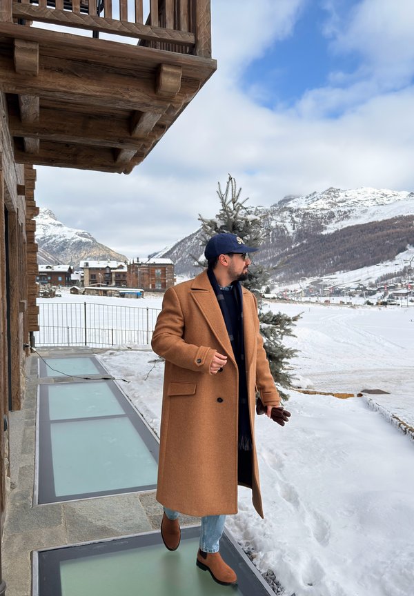 Mann in braunem Mantel, blauer Mütze und Stiefeln, der auf einem verschneiten Weg neben einem Holzgebäude mit schneebedeckten Bergen im Hintergrund geht.