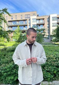 Camicia button-up beige chiaro con una finitura texture e una tasca, indossata sopra un top scuro. Anelli argento e oro sulle mani. Sfondo di fogliame verde.