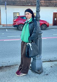 Donna con capelli scuri che indossa una giacca verde, una giacca di pelle nera, pantaloni marroni e sneakers rosse, appoggiata a un lampione.