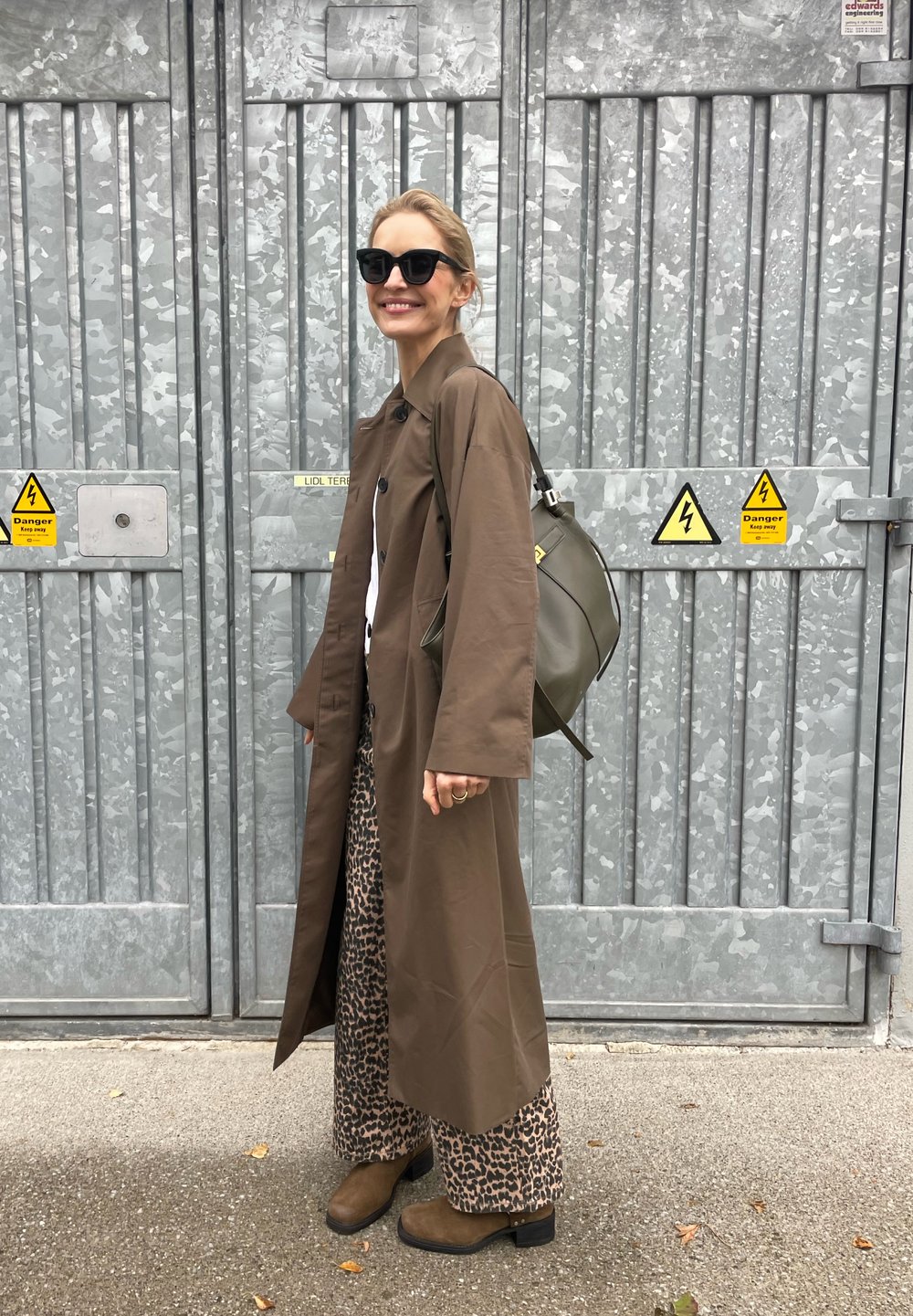 Brown long coat, white shirt, and leopard-print trousers. Brown ankle boots and olive green bag. Black sunglasses. Grey metallic background.