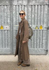 Brown long coat, white shirt, and leopard-print trousers. Brown ankle boots and olive green bag. Black sunglasses. Grey metallic background.