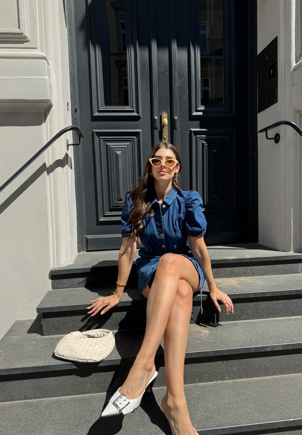 Džinsinė suknelė su pūstomis rankovėmis ir mygtukais, derinama su baltomis aukštakulnėmis šlepet�ėmis. Aksesuarai – geltonos katės akies akiniai nuo saulės ir tekstūruotas baltas maišelis.