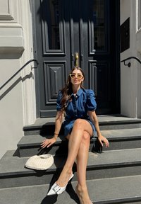 Džinsinė suknelė su pūstomis rankovėmis ir mygtukais, derinama su baltomis aukštakulnėmis šlepetėmis. Aksesuarai – geltonos katės akies akiniai nuo saulės ir tekstūruotas baltas maišelis.