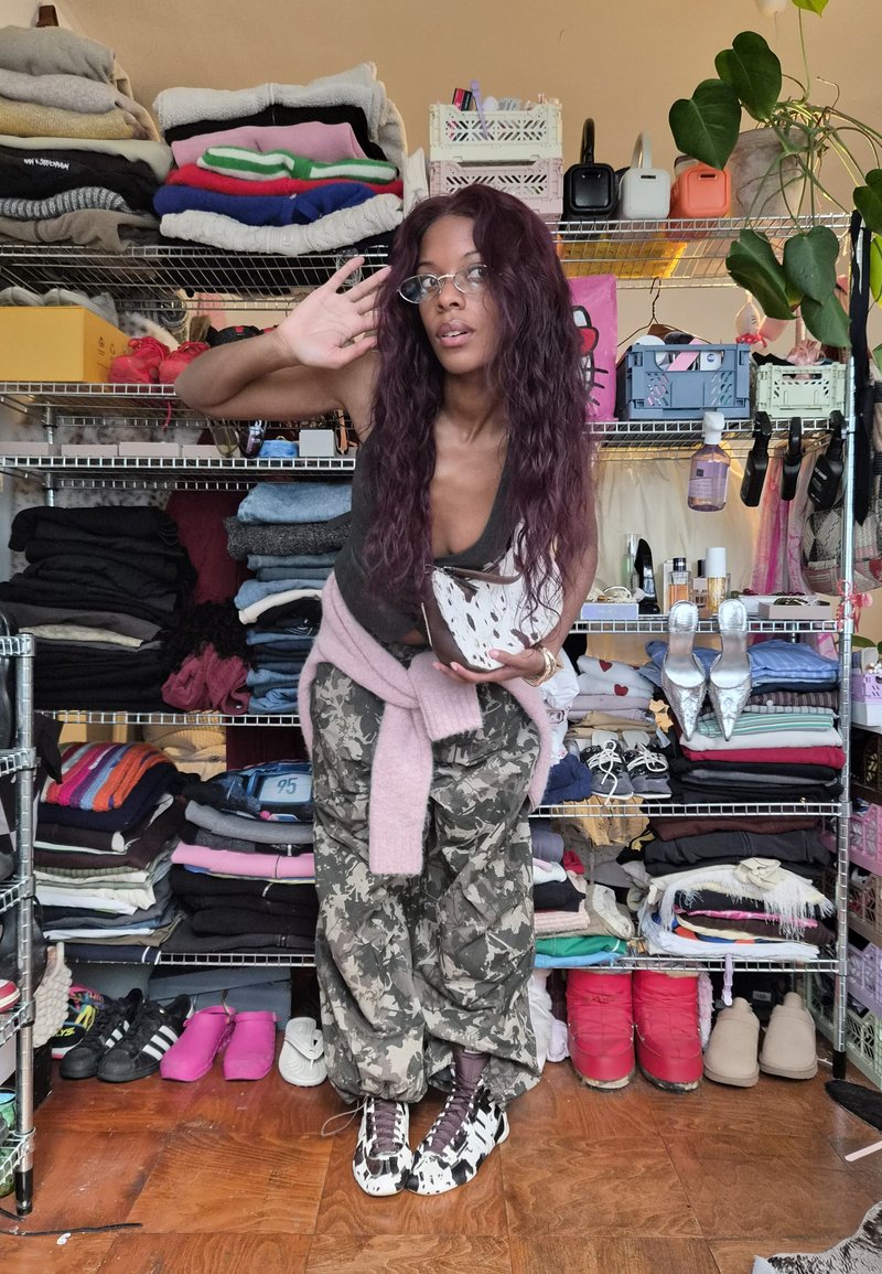 Camo-patterned pants, gray tank top, and light pink sweater tied around waist. Black and white patterned shoes. Shelves with folded clothes.