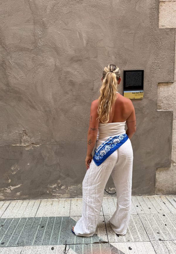 White linen ensemble with wide-leg trousers, accented by a blue patterned sash adorned with white swirls. The background features a textured grey wall.