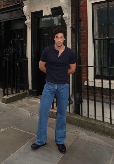 Camiseta henley de manga corta azul oscuro, vaqueros acampanados azul claro y zapatos de cuero negros. De pie frente a una pared de ladrillos con puertas al fondo.