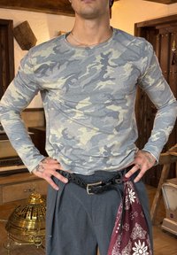 Langærmet grå camouflage skjorte med creme detaljer, tætsiddende design. Bæres sammen med mørke bukser, et sort bælte og et bordeaux mønstret bandana.