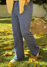 Άτομο που φοράει μπλε καρό παντελόνι, κίτρινο πουκάμισο και λευκά παπούτσια, στέκεται πάνω σε γρασίδι με σκορπισμένα κίτρινα φθινοπωρινά φύλλα.