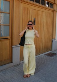 Top corto a righe gialle e pantaloni larghi abbinati, indossati con sandali flip-flop marroni e una borsa a tracolla nera. Sfondo in legno.