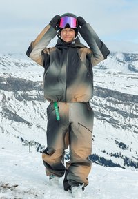 Άτομο που φοράει καφέ και μαύρη στολή σκι ρυθμίζει μαύρο κράνος με μοβ γυαλιά, στέκεται σε χιονισμένο βουνό με χιονισμένους βράχους στο φόντο.