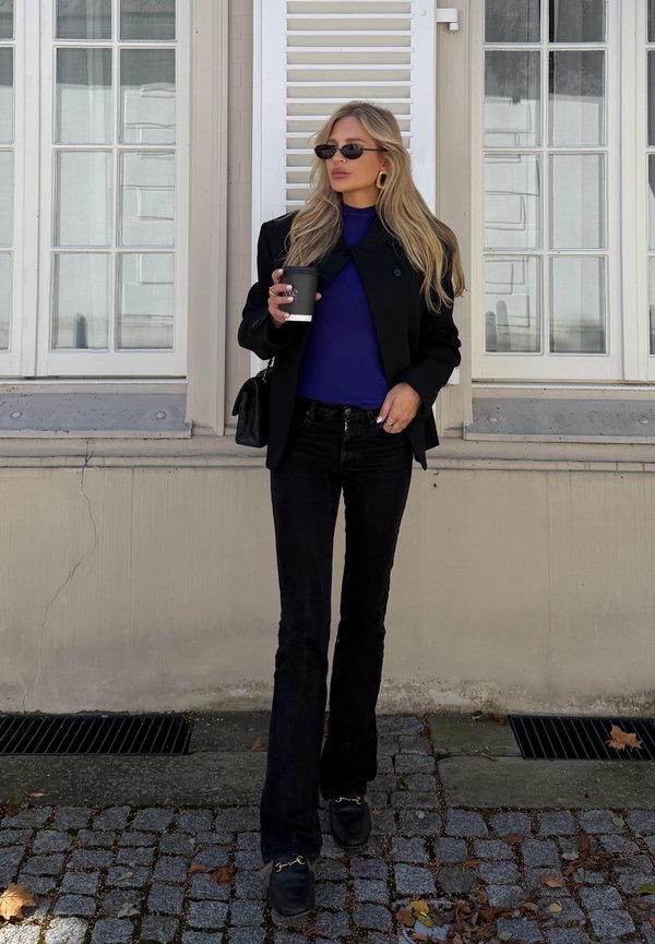 Mujer con cabello largo rubio, con gafas de sol, blazer negro, top morado, pantalones negros, sosteniendo una taza de café, de pie en una calle empedrada.