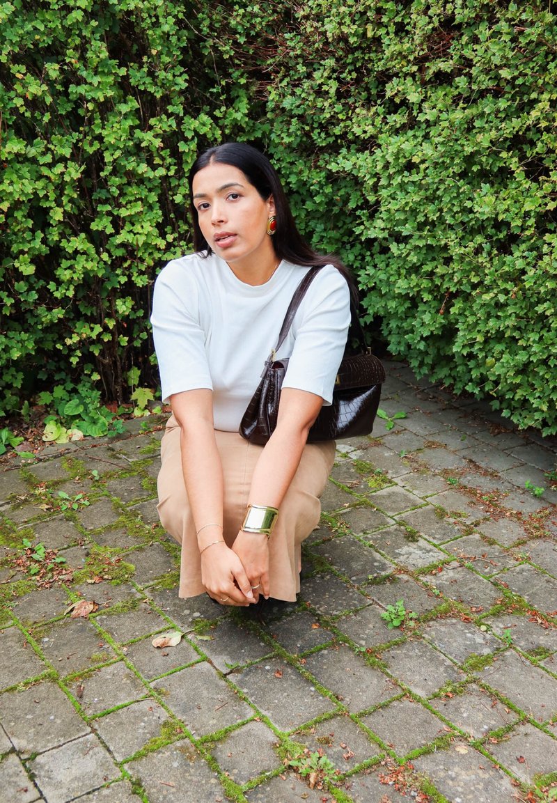 Mujer con cabello largo y oscuro, con camiseta blanca y falda beige, agachada sobre un pavimento de piedra junto a arbustos verdes densos, sosteniendo las manos cerca de los pies.