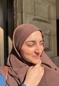 Un hijab in tessuto marrone si adagia attorno alla testa e al collo; presenta una texture liscia. La mano del soggetto tocca il mento, indossando una maglia blu.