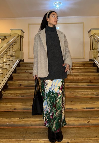 Mujer con abrigo gris y suéter de cuello alto oscuro, sosteniendo un bolso negro, de pie en una escalera de madera en un ambiente interior tenue.