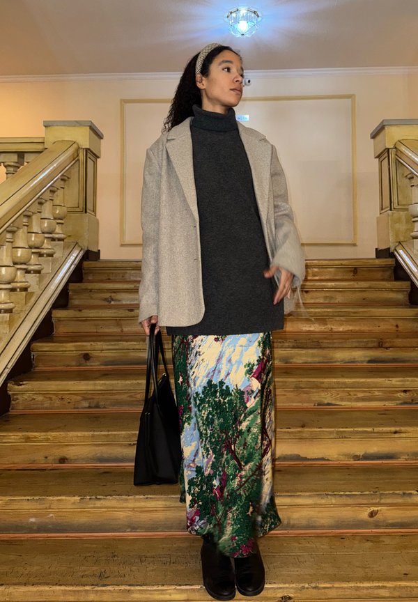 Woman in grey coat and dark roll-neck jumper, holding black bag, standing on wooden stairs in a dim indoor setting.