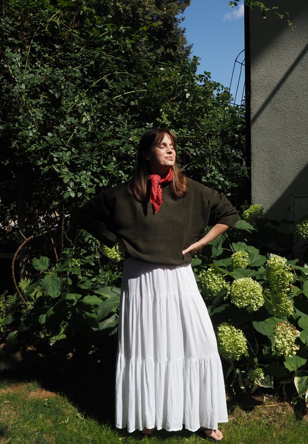 Kvinde stående med lukkede øjne i sollys iført mørkegrøn sweater, rød tørklæde og langt hvidt skørt i en have med grønne planter.