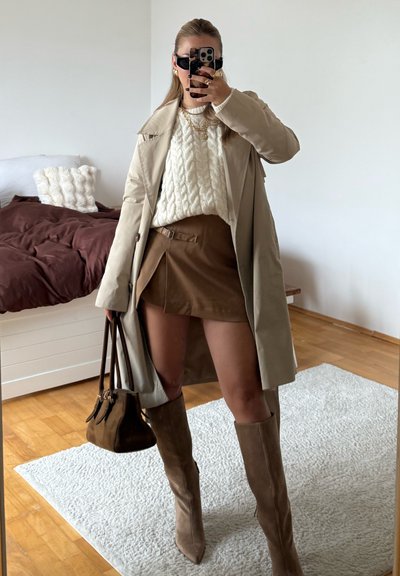 Abrigo trench beige, suéter de punto crema, falda corta marrón con cinturón, botas altas color crema, sosteniendo un bolso marrón, de pie sobre una alfombra blanca.