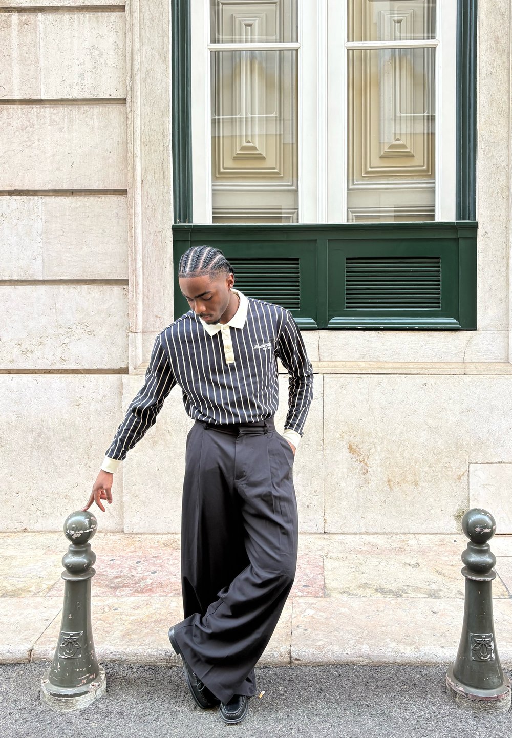 Czarna i biała w paski koszula z długim rękawem z kremowym kołnierzykiem, w połączeniu z wysokimi, czarnymi spodniami z szerokimi nogawkami, stojąca obok słupa ulicznego.