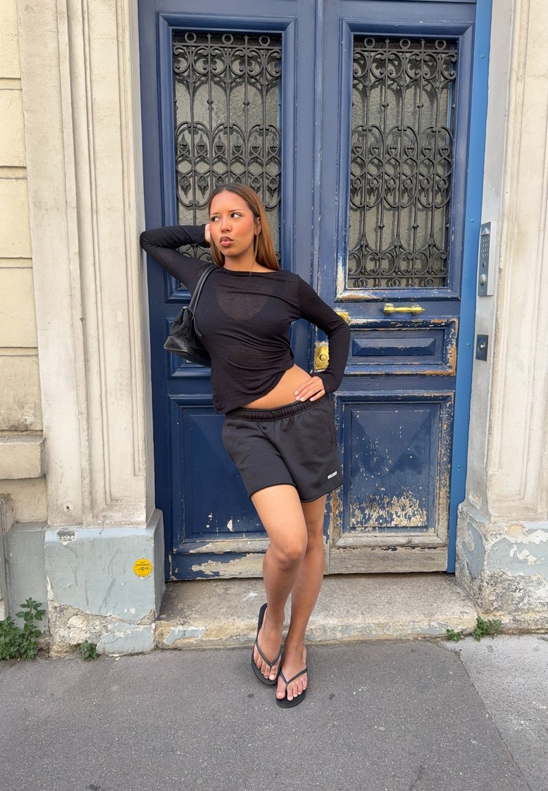 Jeune femme en haut noir, short et sandales pose avec une main sur la hanche et l'autre derrière la tête près d'une porte bleue et usée.