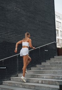 Weißer Sport-BH und Shorts, strukturierte Steinstufen, Metallhandlauf und dunkle Ziegelmauer im Hintergrund. Person steigt die Treppe hinauf.