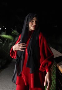 Piros, oversized pulóver bő ujjakkal és passzoló nadrággal; egy laza fekete hidzsábbal párosítva. Az alacsony fényviszonyok kiemelik a színeket.