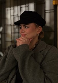 Sivý nadrozmerný kabát so širokými rukávmi, čierna plochá čiapka a zlatý reťazový náhrdelník. Osoba má čistú pokožku a viditeľné piercingy, ruka na brade.