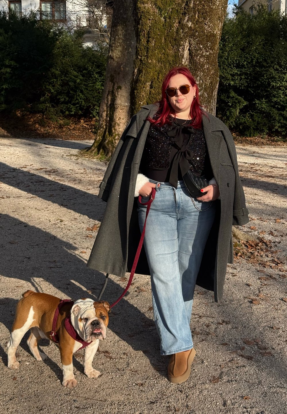 Bouledogue en laisse à côté d'une personne portant un long manteau gris, un haut noir avec un détail en forme de nœud, un jean bleu clair et des bottes marron dans un parc.