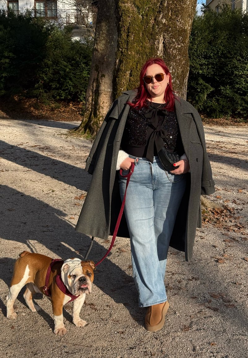 Bouledogue en laisse à côté d'une personne portant un long manteau gris et un haut noir avec un détail en nœud, un jean bleu clair et des bottes marron dans un parc.