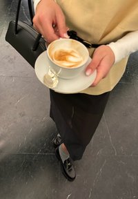 White ceramic cup with latte art, held in hand, paired with a small silver spoon. Black handbag, grey socks, and black loafers visible.