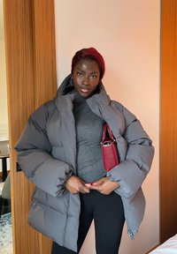 Femme à la peau foncée et aux cheveux rouges portant une doudoune grise par-dessus un haut gris et un pantalon noir, tenant un sac à main rouge à l'intérieur.