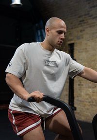 Grå t-skjorte med grafisk trykk og røde treningsshorts med hvite detaljer; personen trener på en maskin med svarte håndtak.