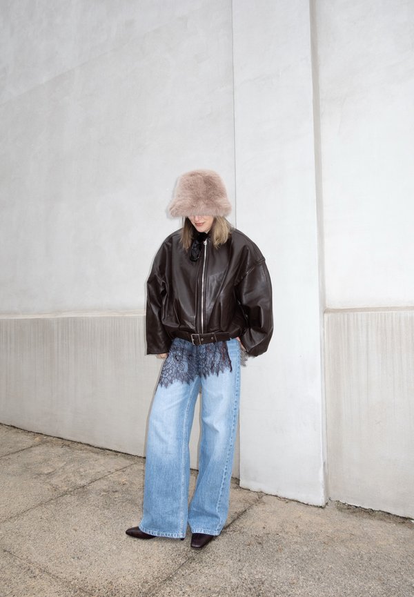 Person, die eine übergroße braune Lederjacke, weit geschnittene hellblaue Jeans mit Spitzenbesatz, einen flauschigen beigen Hut und dunkle spitze Schuhe trägt, steht an einer Wand.