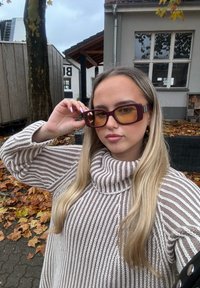 Bruine oversized zonnebril met gele lenzen, vierkante vorm, dikke monturen en zilveren details. Geselecteerd door een persoon in een gestreepte trui.