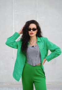 Zelena lanena srajca s gumbi, oblečena čez črno-bele karirane crop top, v kombinaciji z teksturiranimi zelenimi hlačami z visokim pasom. Nosijo se temna sončna očala.