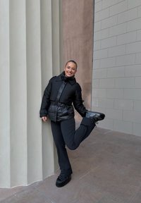 Schwarze Steppjacke mit Taillengürtel, kombiniert mit schwarzen ausgestellten Hosen und klobigen schwarzen Schuhen. Das Modell posiert mit einem erhobenen Bein.