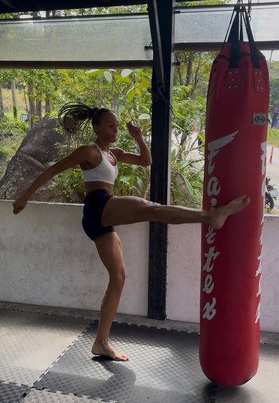 Mujer con ropa deportiva realizando una patada alta en un saco de boxeo rojo en un gimnasio al aire libre con árboles visibles en el exterior.